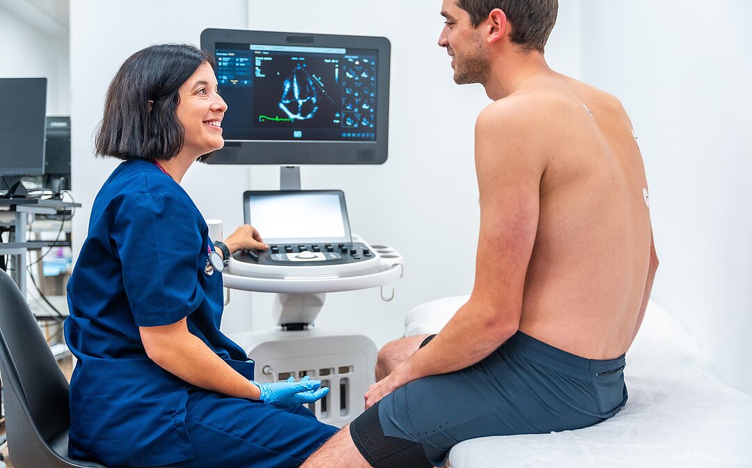 Clinician talks with a male patient seated on an exam table while a heart ultrasound image is displayed on a monitor.